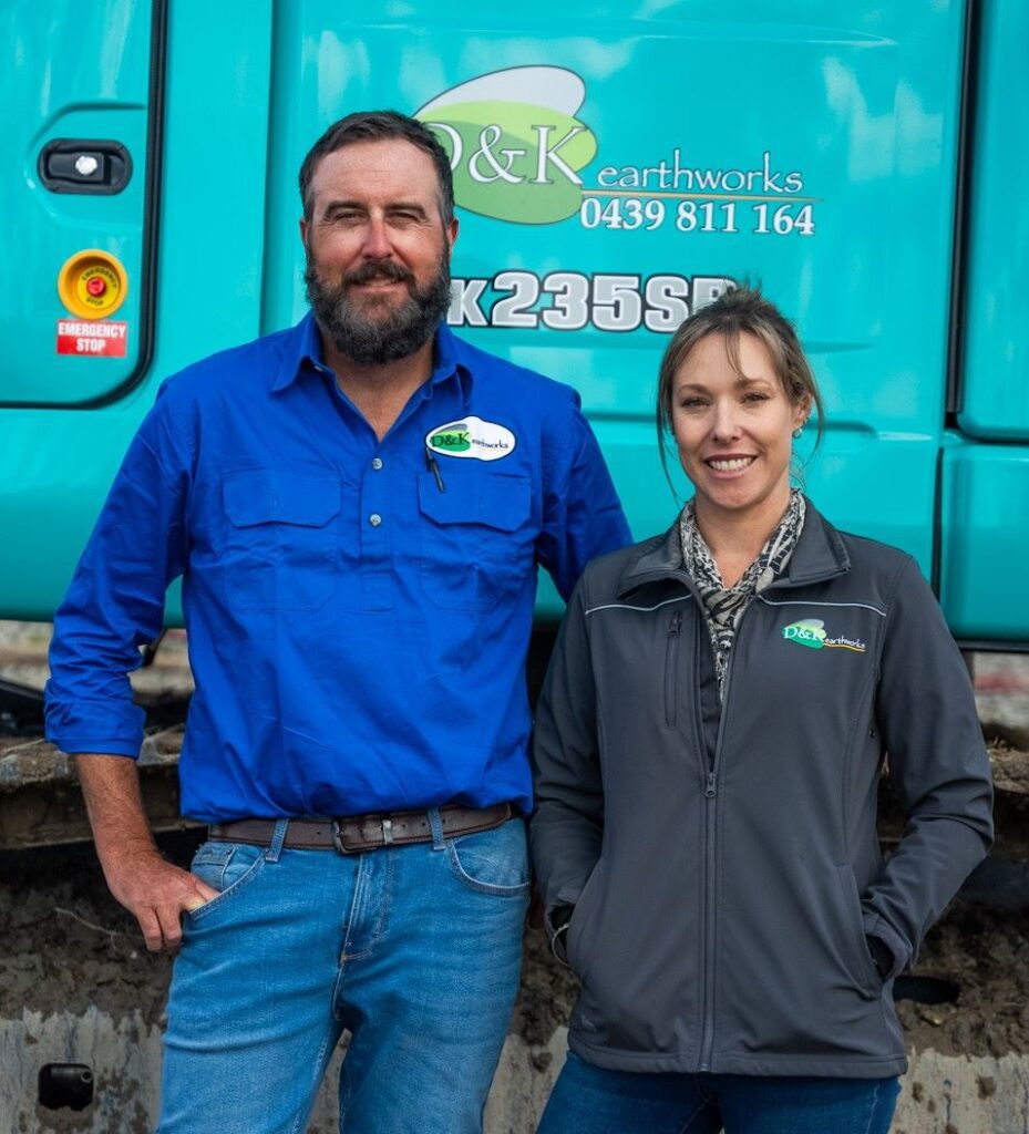A man and a woman in work attire stand in front of a teal R&K Earthworks vehicle. Both are smiling and appear to be on a construction site. The vehicle has contact information and a logo displayed.