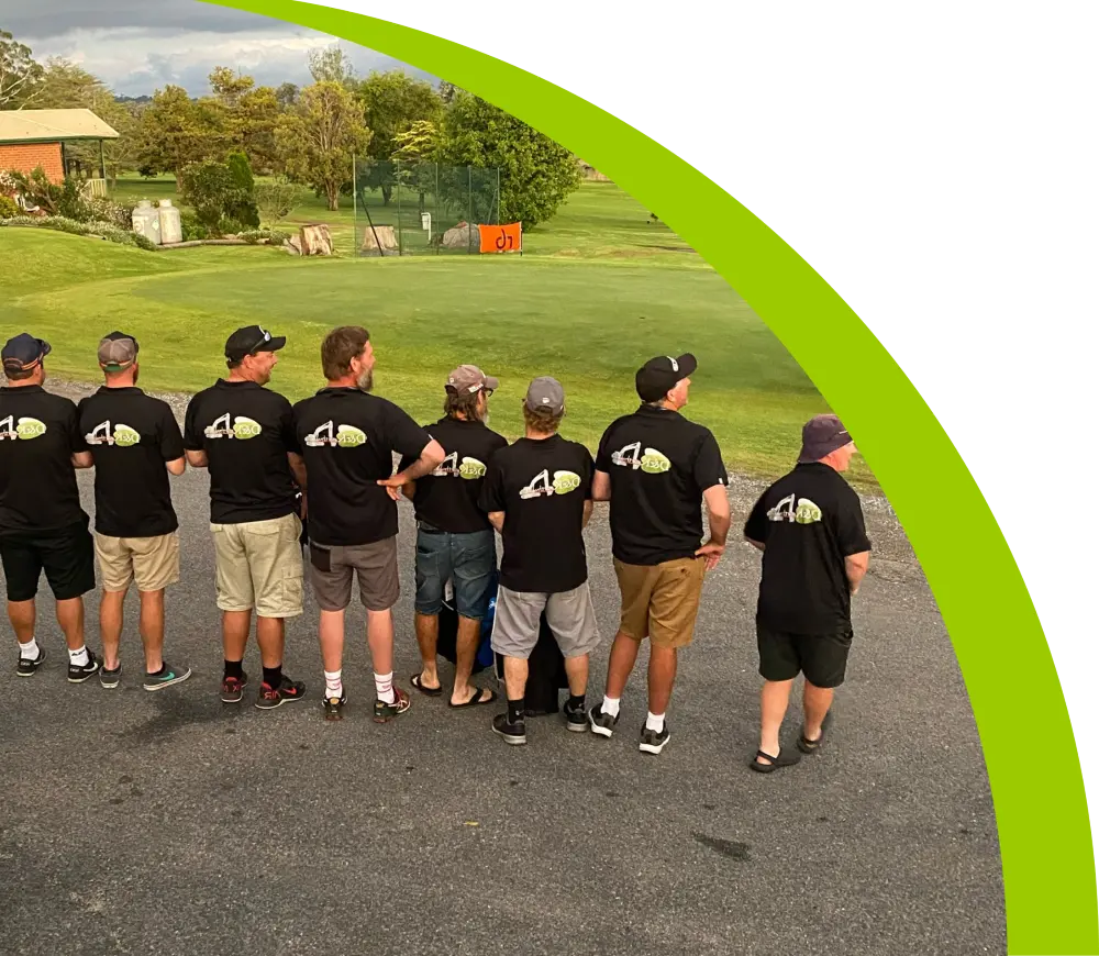 Eight men in matching black T-shirts and caps stand in a row, facing away on a paved area near a green lawn with trees and a house in the background. A curved green stripe partially frames the image.