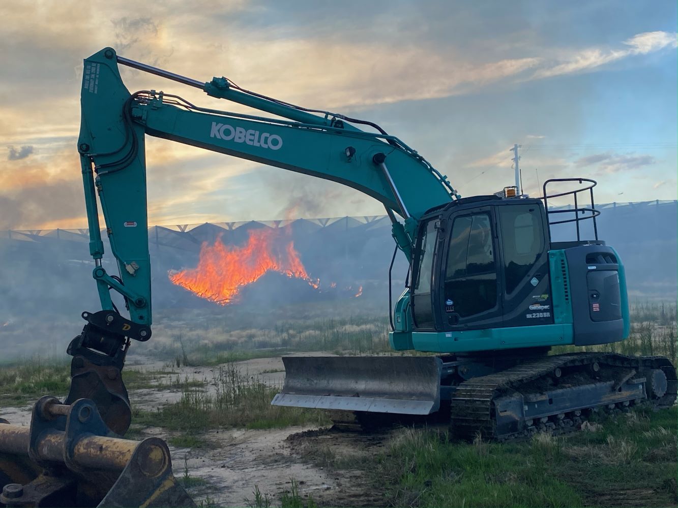 A blue excavator is parked on grassy terrain with its arm extended. In the background, flames and smoke rise against a cloudy sky, suggesting a fire behind a structure. The setting is outdoors, possibly at a construction or industrial site.