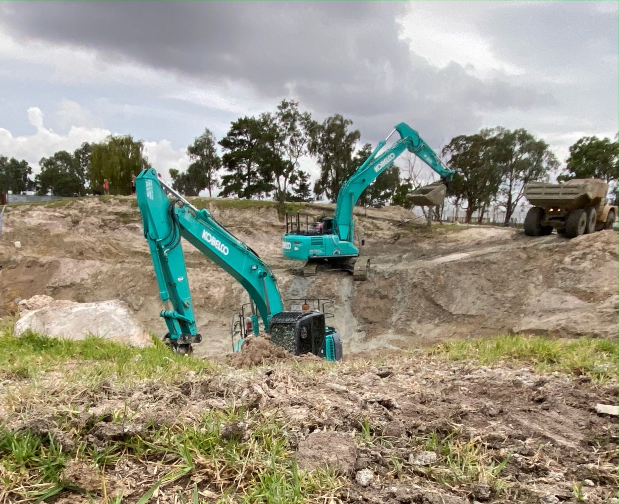 Two blue excavators are working in a large, dirt-filled construction site. In the background, a dump truck is parked near piles of soil. The sky is cloudy, and trees line the horizon.