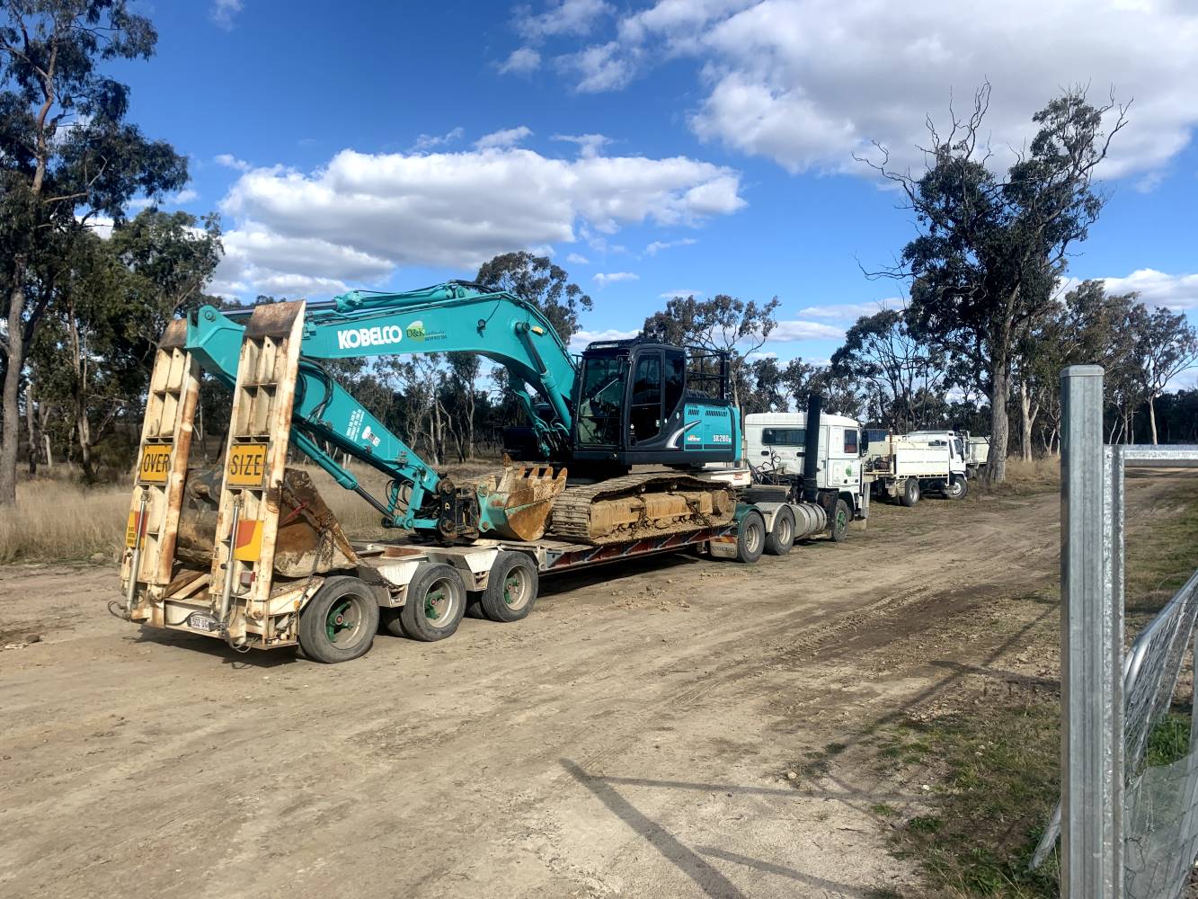 A large green excavator is being transported on a flatbed trailer pulled by a truck. The scene is outdoors on a dirt path, surrounded by tall trees under a partly cloudy sky.