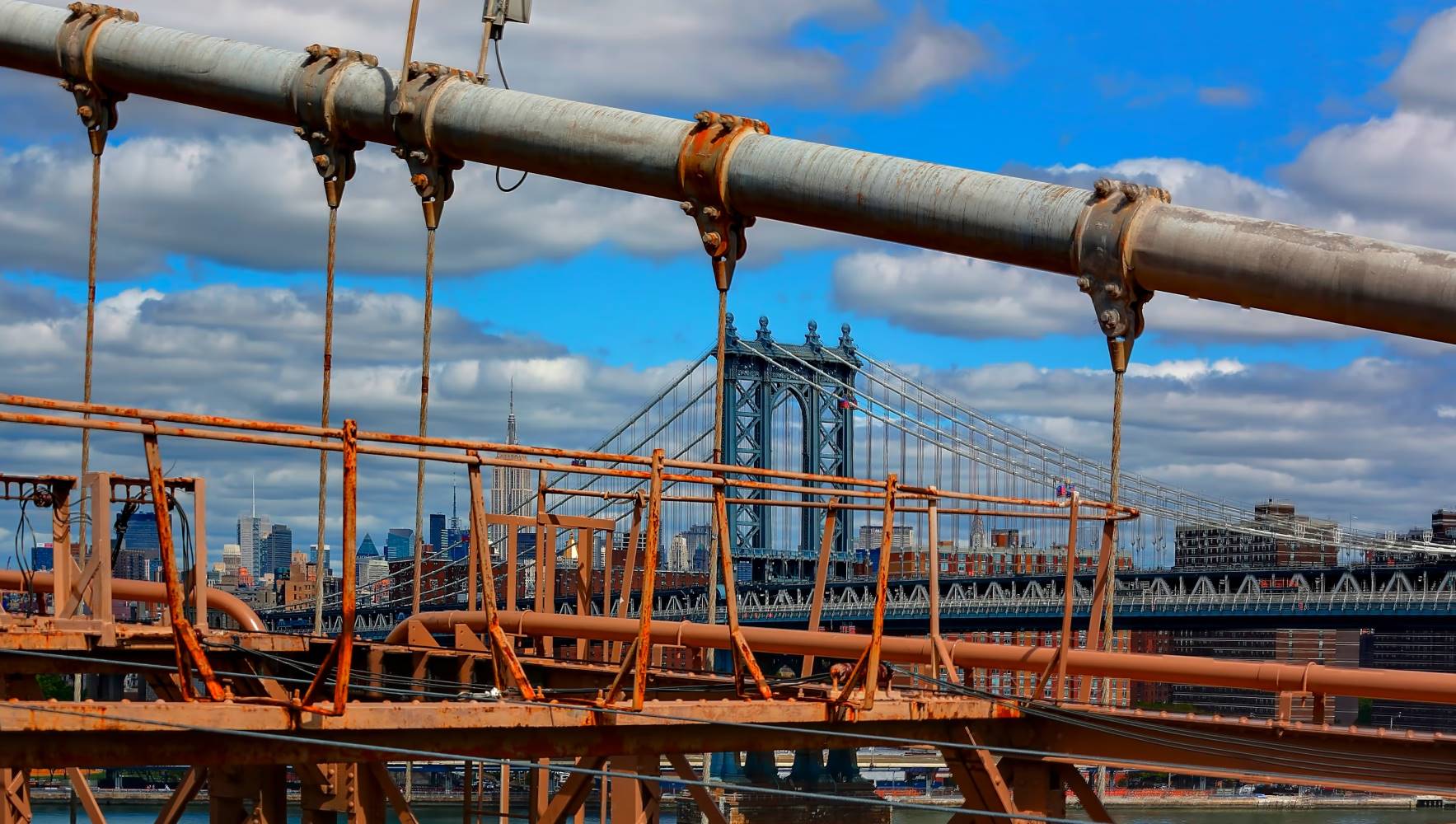 View of the Manhattan Bridge framed by rusted metal beams and pipes, with a city skyline and partly cloudy blue sky in the background.